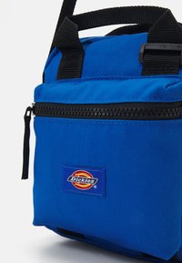 Saco de tecido azul com um bolso frontal com fecho, alças pretas e um emblema. Apresenta uma construção robusta e um design compacto.