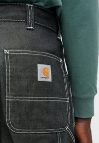 Pantalones de mezclilla negra con un gran bolsillo trasero con costuras blancas y una etiqueta cuadrada de Carhartt en naranja y gris, acompañados de una camisa de manga larga verde.