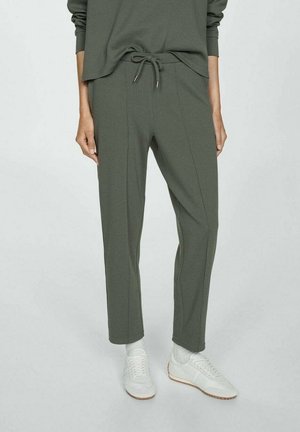 Persoon die olijfgroene broek met trekkoorden draagt, bijpassende top met lange mouwen, witte sokken en witte sneakers, staand tegen een effen achtergrond.