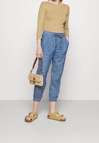 Světle hnědý top s dlouhým rukávem, modré cropped kalhoty z chambray, béžové nazouvací sandály a malá tkaná kabelka s pleteným popruhem.