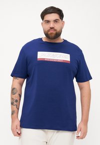 Marineblå t-skjorte med korte ermer laget av bomull, med et grafisk trykk i hvitt og rødt med 'JACK & JONES' og 'SINCE NINETEEN NINETY.'
