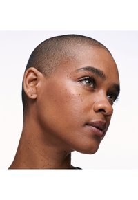 Gros plan d'une personne aux cheveux rasés, peau brune et yeux noisette regardant légèrement vers le haut sur un fond uni et clair.