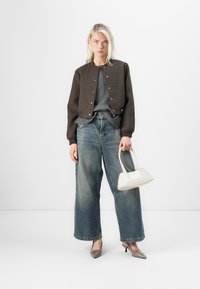 Veste courte marron avec des boutons-pression argentés, haut gris, jean bleu large et sac à main blanc. Le mannequin porte des escarpins gris pointus et a les cheveux blonds raides.
