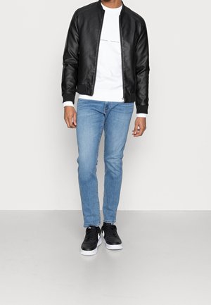 Mies, jolla on musta nahkatakki, valkoinen collegepaita, vaaleansiniset farkut ja mustat lenkkarit, seisoo yksinkertaista vaaleaa taustaa vasten.