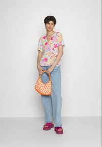 Blusa floral de manga curta com padrões em rosa, amarelo e verde, combinada com jeans azul claros. Segurando uma mala tecida laranja e usando sandálias rosa de plataforma.
