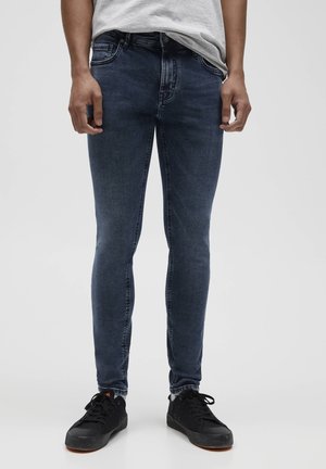 Homme portant un jean slim bleu foncé, un t-shirt gris clair et des baskets noires à lacets, debout devant un fond gris uni.