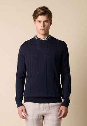 Junger Mann, der einen dunkelblauen Pullover über einem gestreiften Hemd mit Kragen und hellbeiger Hose trägt, vor einem einfarbigen beigen Hintergrund stehend.