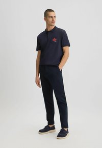 Camisa polo de navy con un bolsillo y un logo rojo, combinada con pantalones oscuros y zapatos navy con suelas color crema. Diseño simple y limpio.