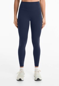 Leggings taille haute bleu marine en matière extensible. Présentent une texture lisse et un design ajusté, associés à des chaussures de sport de couleur claire.