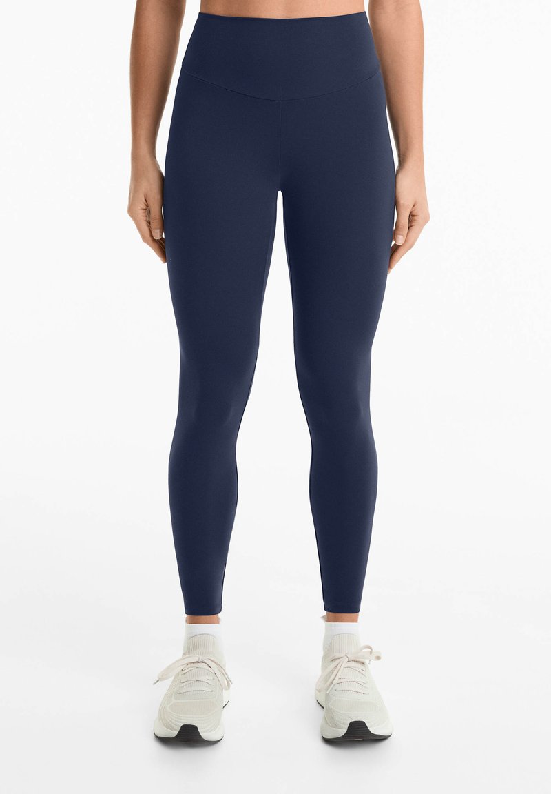 Leggings taille haute bleu marine en matière extensible. Présentent une texture lisse et un design ajusté, associés à des chaussures de sport de couleur claire.
