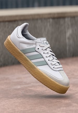 Hvid Adidas Samba sneakers med lysegrønne striber og gummisål svæver over en lyserød betonoverflade.