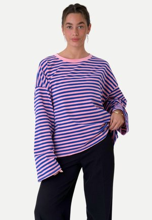 Jeune femme portant une chemise oversize à manches longues rayée rose et bleue avec un pantalon noir, debout avec une main dans la poche sur un fond uni.