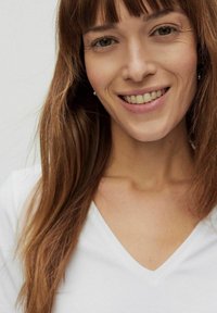 Hvid V-hals t-shirt, glat stof. Modellen har langt, glat brunt hår og et subtilt smil, med hendes træk klart synlige.
