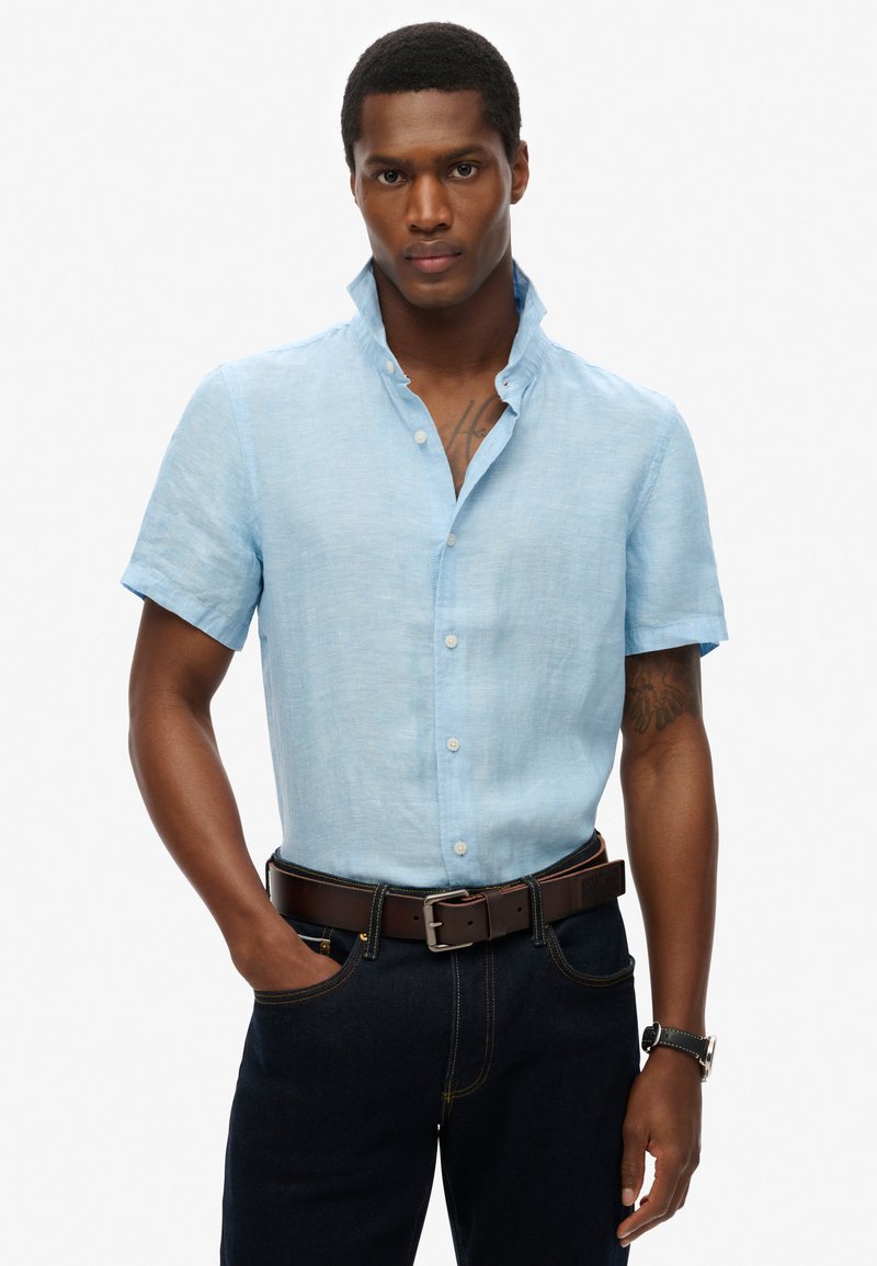 Camicia azzurra a maniche corte realizzata in un tessuto testurizzato, con colletto con bottoni e vestibilità dritta, abbinata a jeans scuri e una cintura marrone.