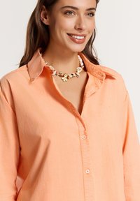 Camicia button-up color pesca con colletto. Indossata con una collana multicolore con accenti dorati ed elementi di design floreale.