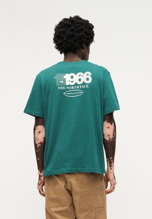 Persona con cabello rizado oscuro y vitíligo en los brazos, vistiendo una camiseta verde azulado de The North Face con el texto "1966" y pantalones cargo beige, de espaldas.
