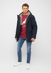 Giacca imbottita blu navy con cappuccio foderato in pelliccia sintetica, abbinata a una felpa bordeaux con grafica di un uccello e jeans blu a taglio stretto, sneakers bianche.