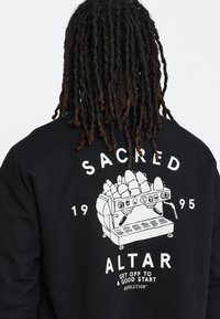 Zwarte sweatshirts met een witte grafiek van een koffiezetapparaat met versieringen. Tekst bevat "SACRED," "ALTAR," en "1995."