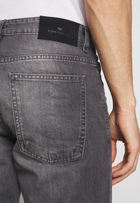 Jeans de mezclilla gris con una etiqueta de cuero oscuro que dice "TOM TAILOR" en la cintura, con bolsillos traseros clásicos y una textura suave.