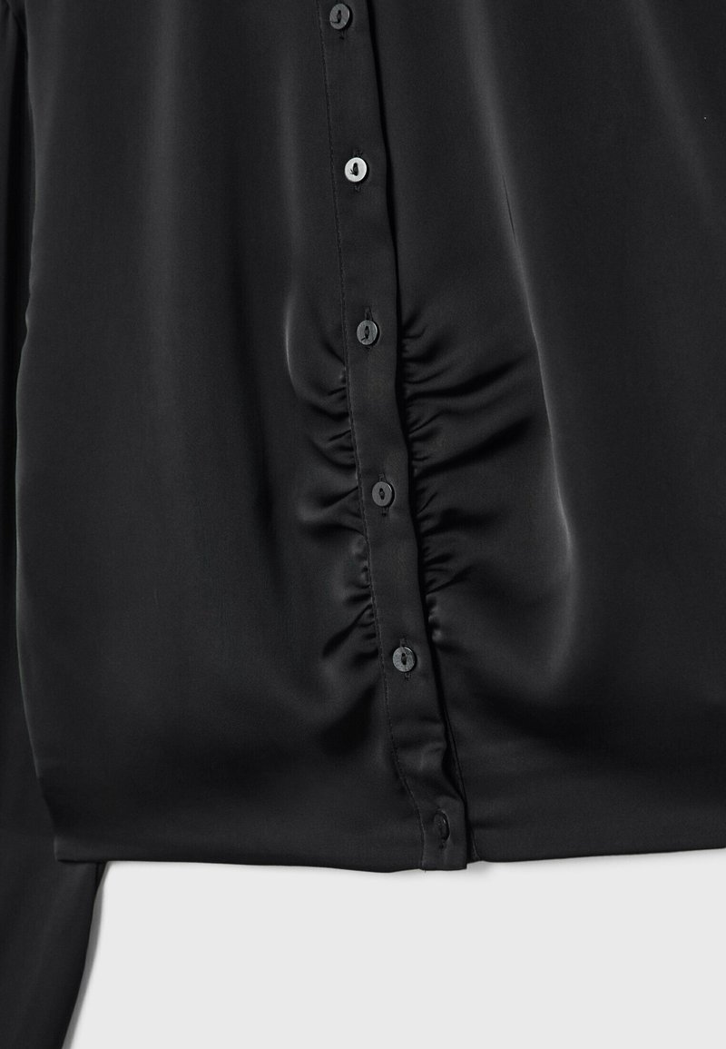 Blouse noire avec une texture en satin lisse, des détails froncés et des boutons noirs le long du devant. Manches longues avec une coupe décontractée.