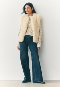 Kreemivärvi faux-fur vest, off-white pitsioberõnge ja tume sinine laiade jalgadega denim teksad. Nähtavad ka mustad jalanõud ja pikad varrukad.