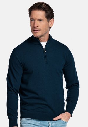 Man draagt een donkerblauwe trui met een rits tot aan de hals over een wit shirt, met lichtblauwe jeans, kijkt opzij tegen een effen achtergrond.