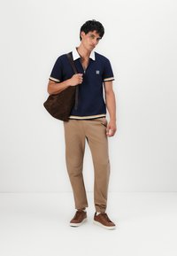 Marineblå polo med hvit krage og stripete detaljer, beige bukser, brune sneakers og en mørkebrun skulderveske.