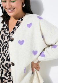 Kremowy kardigan w fioletowe serca, o oversize’owym kroju, z zapięciem na guziki i ściągaczami przy rękawach. Noszony na czarnej sukience w beżowo-czarny zwierzęcy wzór.