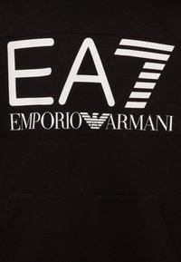 Svart tyg med vit "EA7" text ovanför "Emporio Armani" och varumärkets örnlogotyp i mitten.