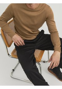 Brown knitted sweater with a round neckline, paired with black pants. Model seated on a woven chair with a chrome frame.