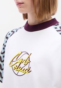 Λευκό μπλουζάκι με μπορντό ριμπ λαιμόκοψη, διακοσμημένο με μανίκια με σχέδιο και κίτρινη κεντητική με την επιγραφή "Carlo Colucci" κ αι γραφικό μπάσκετ.