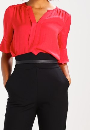 Blouse rouge à manches courtes avec un col en V et des épaules froncées, associée à un pantalon noir taille haute avec une ceinture noire fine.