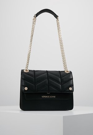 Sac à main en cuir matelassé noir avec bandoulière chaîne dorée et logo "Versace Jeans" sur le rabat avant, présenté sur une surface blanche.