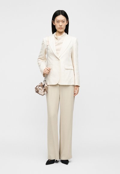 Γυναίκα όρθια, φοράει εκρού μπλέιζερ και παντελόνες με φαρδιά μπατζάκια, κρατώντας μια μικρή μπεζ χρωματιστή τσάντα με ασημένια αλυσίδα.