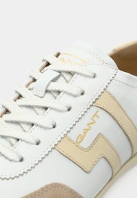 Άσπρο αθλητικό παπούτσι με μπεζ λεπτομέρειες, λευκά κορδόνια και το λογότυπο "GANT" σε χρυσό στη πλευρά.