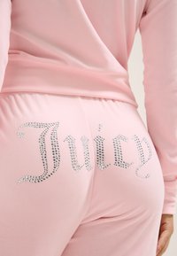 Rožiniai sportiniai trikotažo kelnių, pagamintų iš minkštos medžiagos, su kreivu juosmeniu ir sidabriniais kristalais užrašytu "Juicy" ant užpakalinės kišenės.