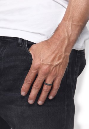 Schwarzer Metallring an einer Hand, die nahe einer Tasche ruht, mit ausgeprägten Venen und hellem Hautton; getragen zu dunklen Jeans und einem weißen Hemd.