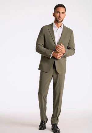 Olijfgroen pak met een gestructureerd ontwerp, enkelvoudige blazer, bijpassende broek en zwarte leren schoenen. Witte shirt eronder.