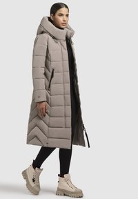 Beež polsterdatud jakk kõrge kraega, millel on tõmblukk ja küljetaskud. Kanda mustade legginsite ja beežide jalatsitega.