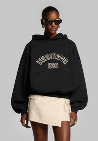 Black oversized hoodie with white "WRSTBHV" text and embroidery, paired with a beige mini skirt. Features a front pocket and soft fabric.