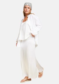 Hvit lin skjorte lagd over en hvit flytende maxikjole med sidespalter, komplementert av et mønstret hodeplagg og beige flip-flops.