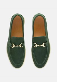 Groene suède instappers met een gouden hardware accent, ronde neuzen en een contrasterende crèmekleurige rubberen zool. Geen zichtbare patronen of texturen.