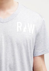 Světle šedé tričko s výstřihem do V vyrobené z měkké, texturované látky. Obsahuje bílé logo s nápisem "RAW" a menším textem níže. Má krátké rukávy.