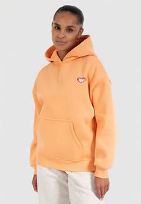 Oranje hoodie gemaakt van zacht materiaal, voorzien van een voorkantzak en een klein geborduurd logo op de linkerborst. Ontworpen met een capuchon met trekkoorden.