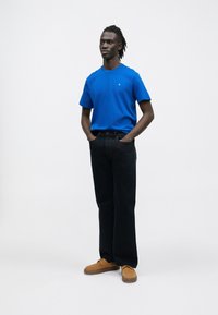 Niebieska bawełniana koszulka, czarne spodnie dżinsowe i brązowe zamszowe buty. Model stoi z rękami w kieszeniach na jednolitym białym tle.