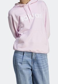 Rosa hoodie med en stor vit "adidas"-logotyp, med en framficka och avslappnad passform, matchad med ljusblå jeans.