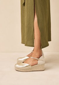 Metalliska guld-espadriller med kilklack i texturerat tyg, med en spännesrem runt ankeln och plattform klädd i jute, matchade med en olivfärgad klänning.