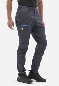 Ciemnoniebieskie dżinsy z kontrastowymi szwami, z zamykaną na zamek kieszenią w niebieskim kolorze. Noszone z czarnymi butami turystycznymi. Regulowany pasek w talii.