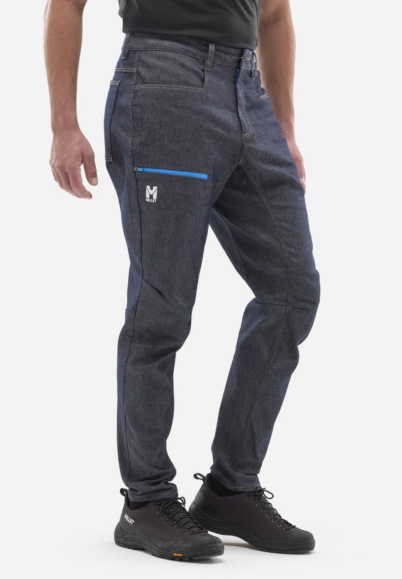Ciemnoniebieskie dżinsy z kontrastowymi szwami, z zamykaną na zamek kieszenią w niebieskim kolorze. Noszone z czarnymi butami turystycznymi. Regulowany pasek w talii.