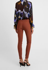 Leggings ajustés marron associés à un chemisier noir transparent avec un motif floral coloré et des manches volantes, portés avec des sandales à talons noires.
